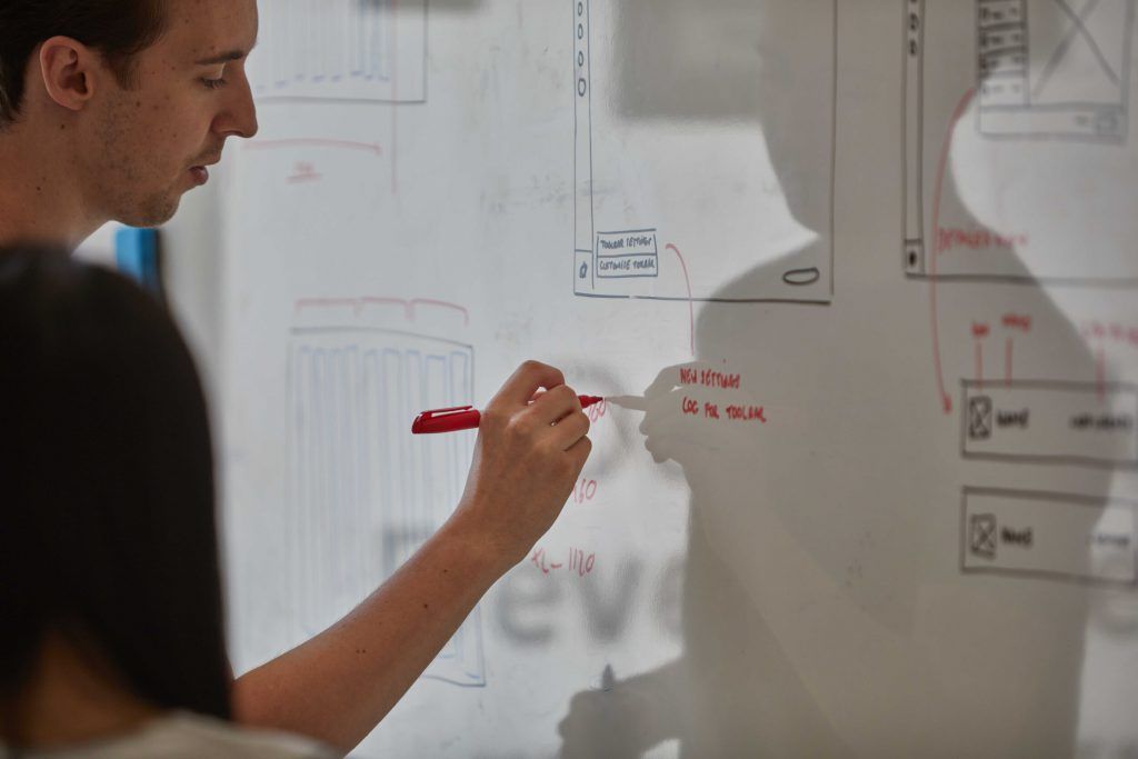 Two people at a whiteboard sketching app screens, one annotating a toolbar in red pen