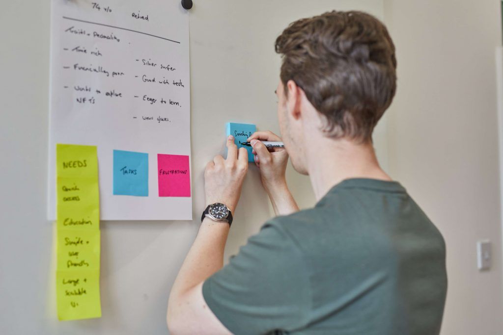 Man writing a sticky note beside a flipchart persona for a 74 year old retired user, with columns for needs, tasks and frustrations