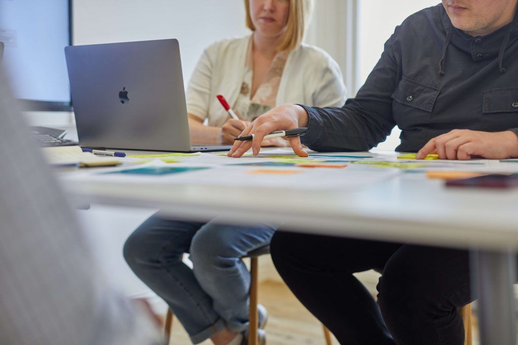 Two people at a table working through printed screens and coloured sticky notes with a laptop open