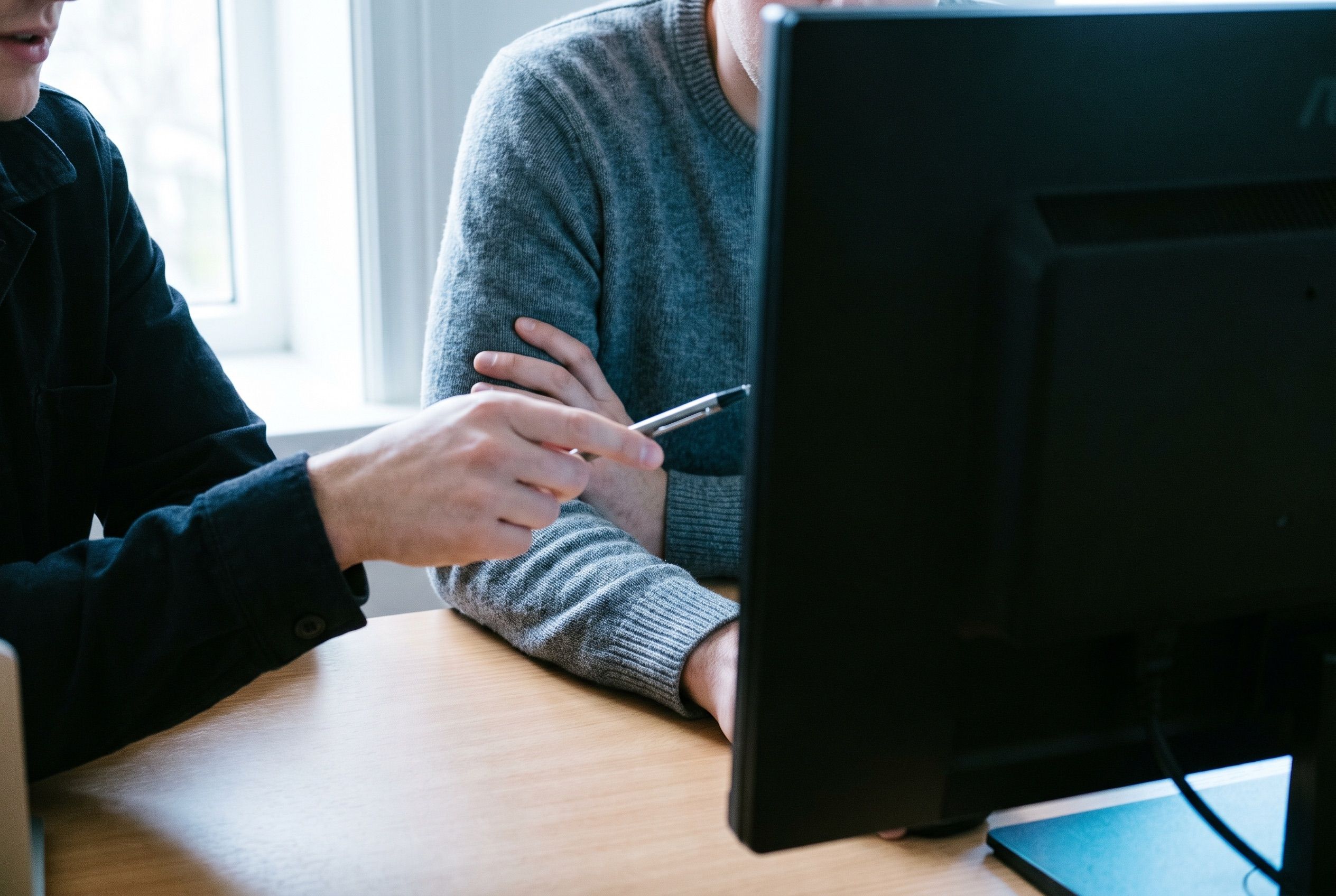 Two people at a desk, one pointing a pen at a monitor turned away from the camera.