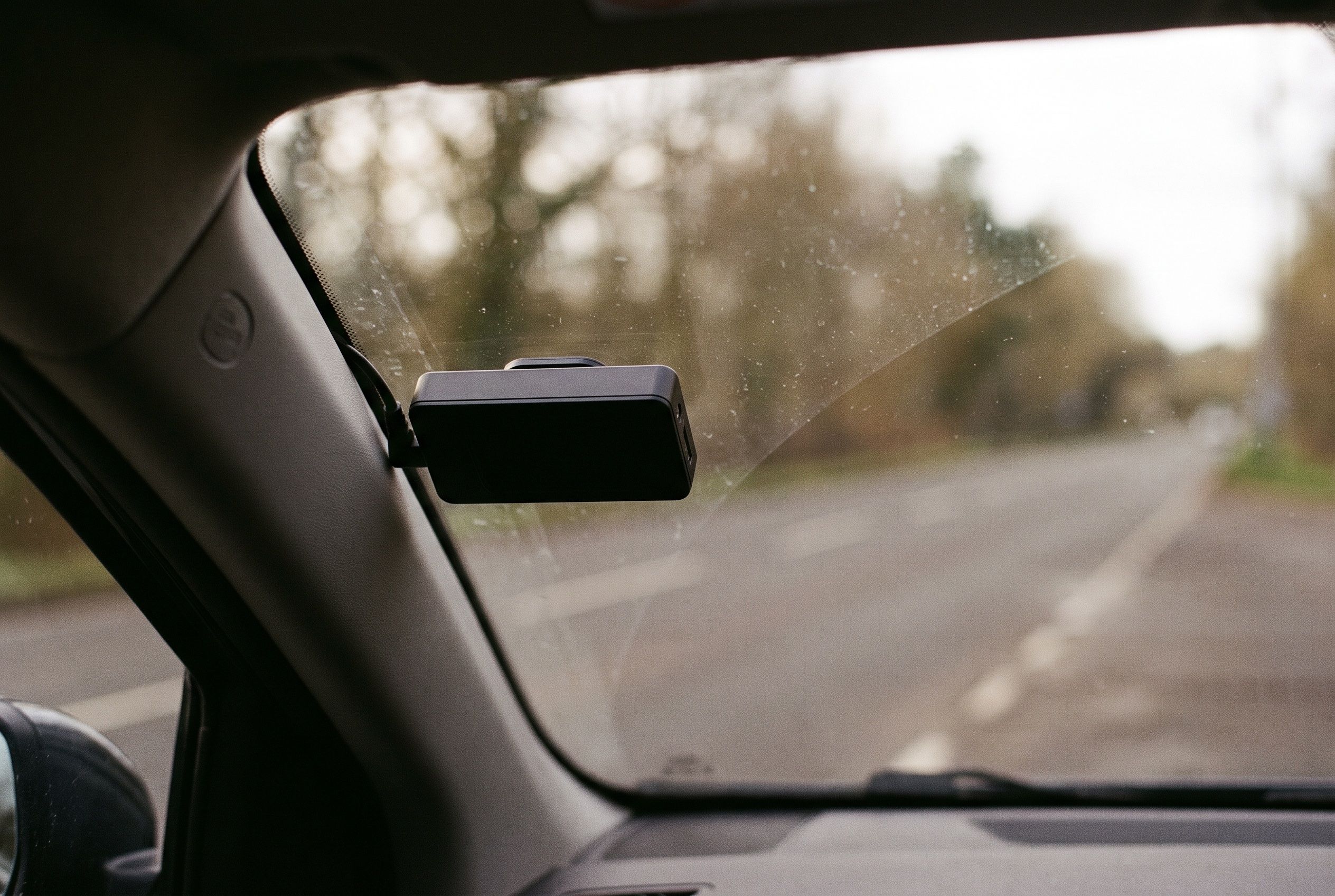 A compact in-car telematics device mounted at the top of a car windscreen, the road ahead beyond it.