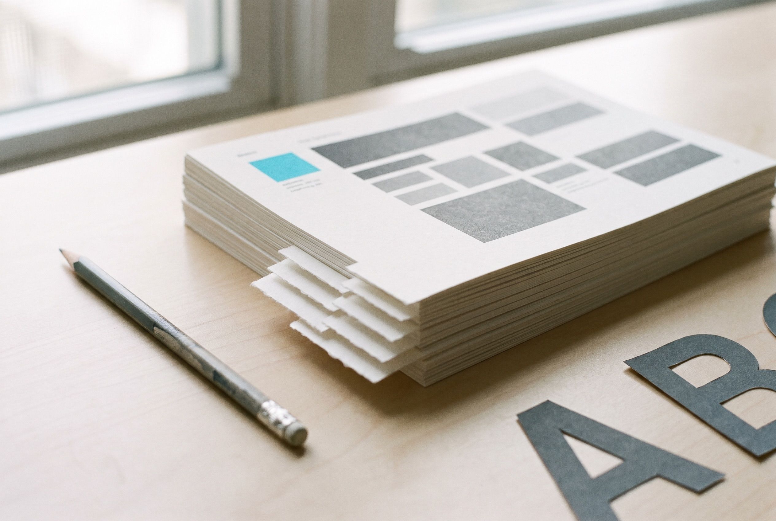 A stack of printed brand guideline pages with a single teal swatch on a pale wooden desk, a pencil alongside and large cut-paper letterforms at the edge of the frame.