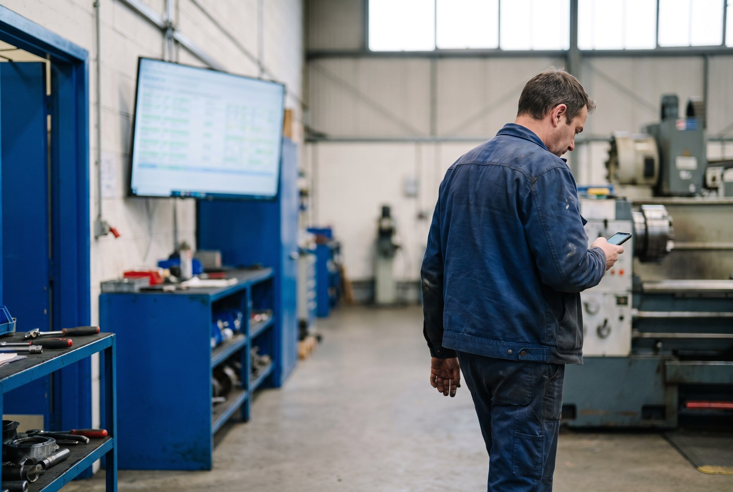 A maintenance engineer on a workshop floor reading something on his phone, a wall-mounted dashboard screen behind him unattended.