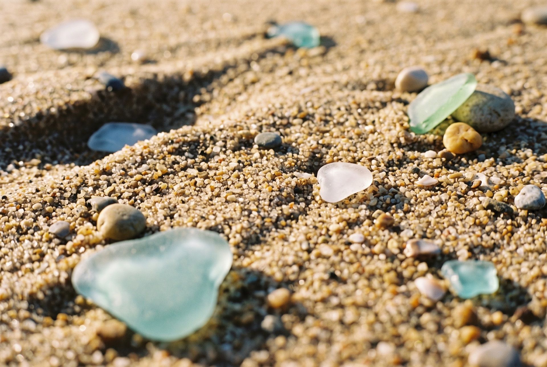 Coarse golden sand at the tide line with seven pieces of frosted sea glass, aqua, white and green, scattered among small pebbles.