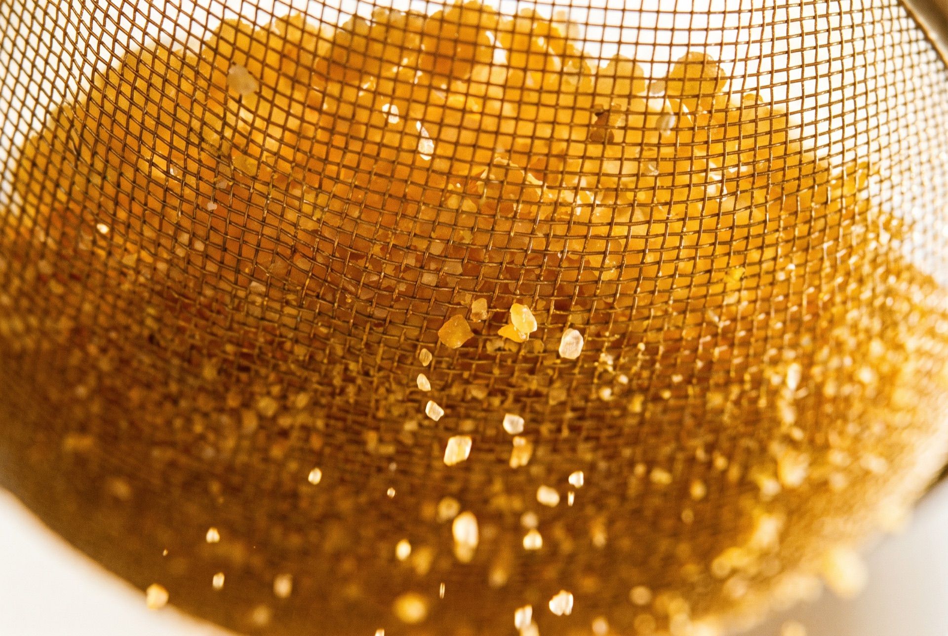 A close-up of a fine brass mesh sieve holding coarse amber grains, with a few smaller grains falling through the mesh.