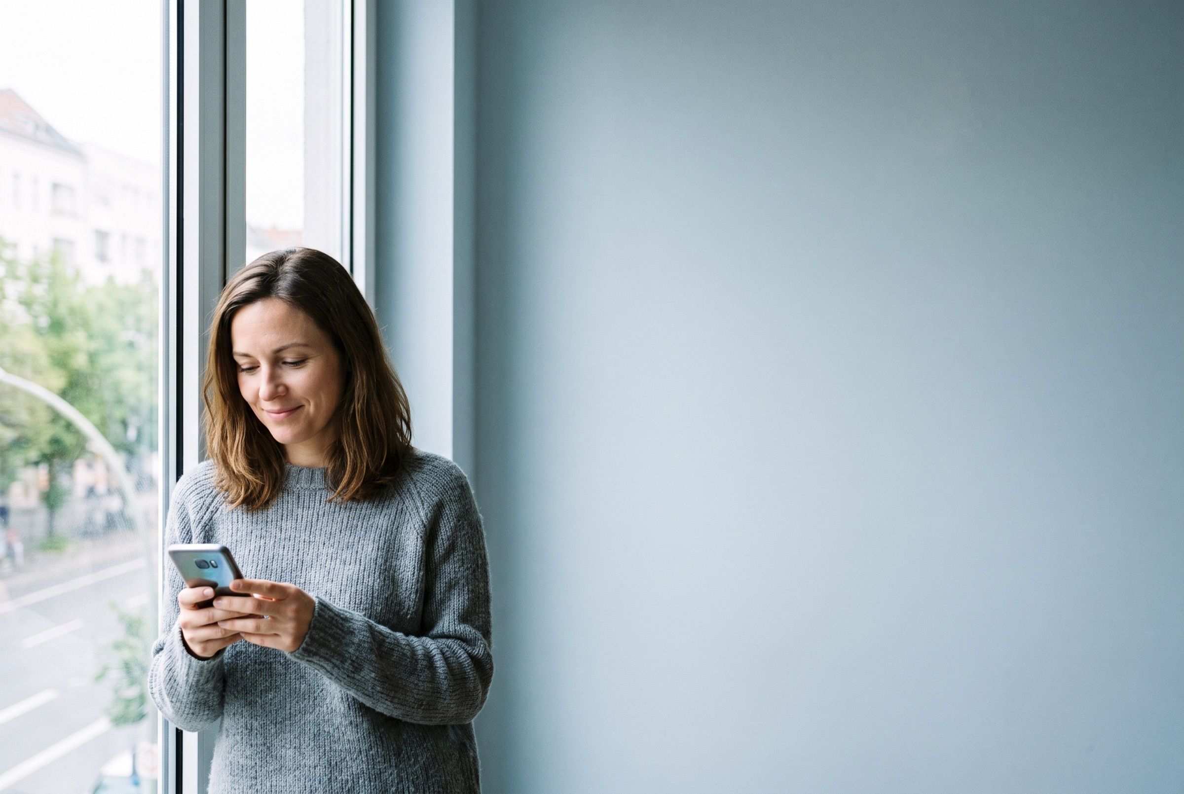 A woman standing by a window, calmly checking a banking app on her phone.