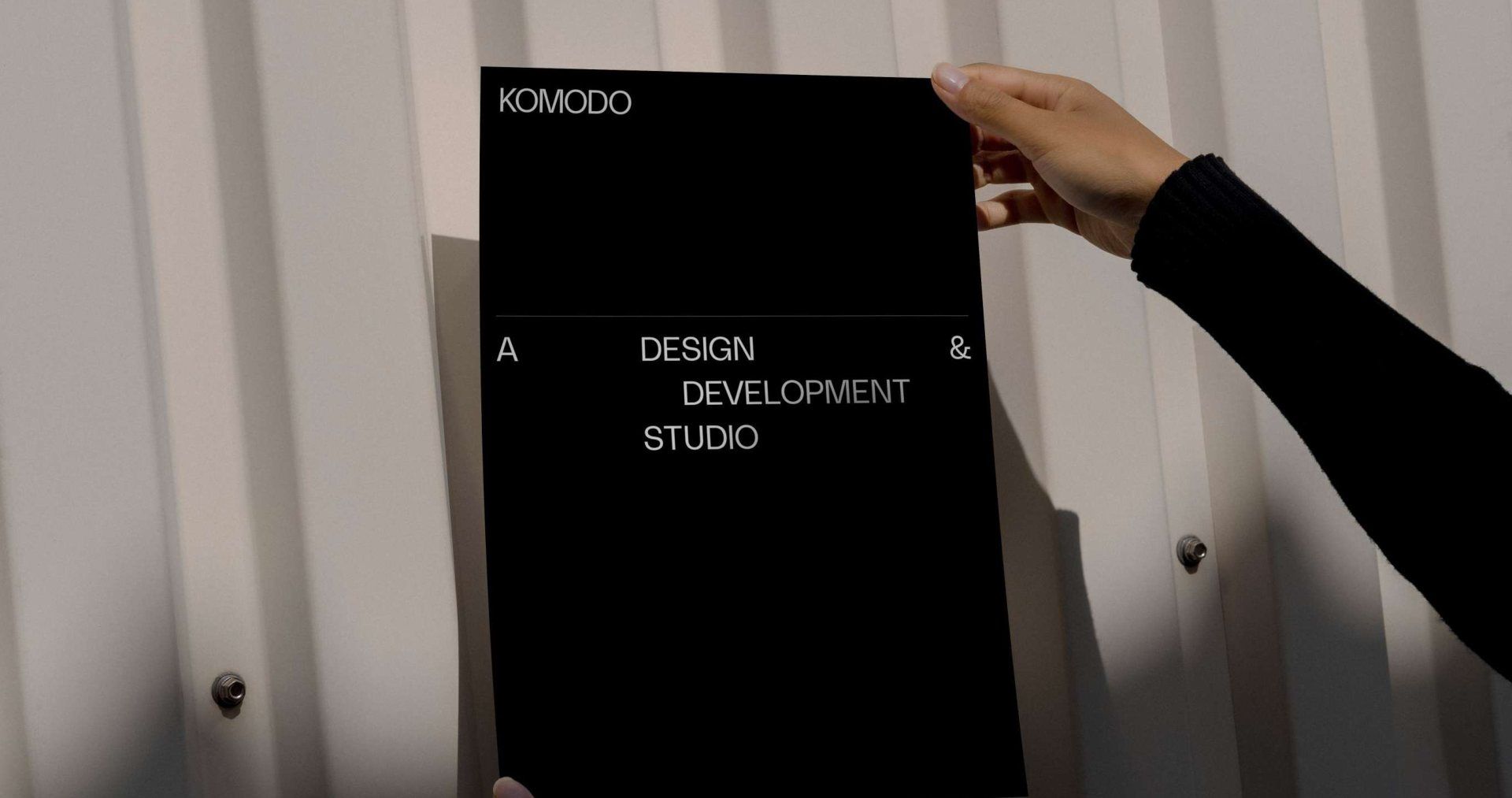 A hand holding a black Komodo poster against a corrugated metal wall, the words A DESIGN AND DEVELOPMENT STUDIO stepped across it line by line.
