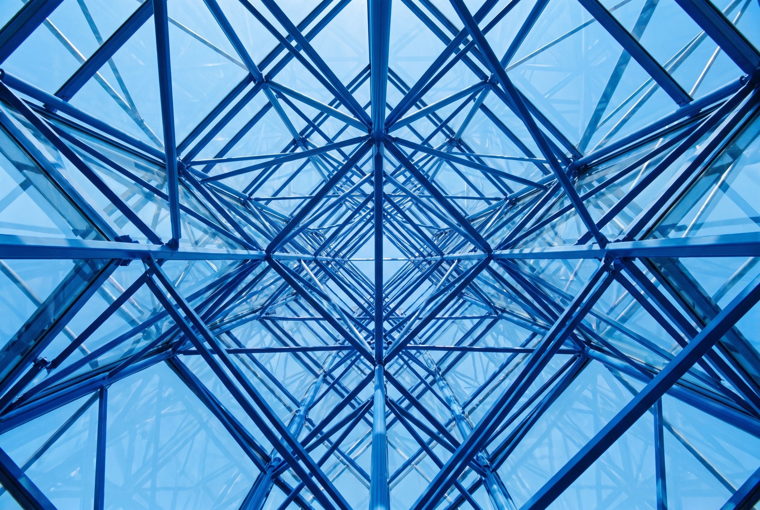 Looking up into a cobalt-blue lattice of steel and glass, a precise geometric structure against bright sky.