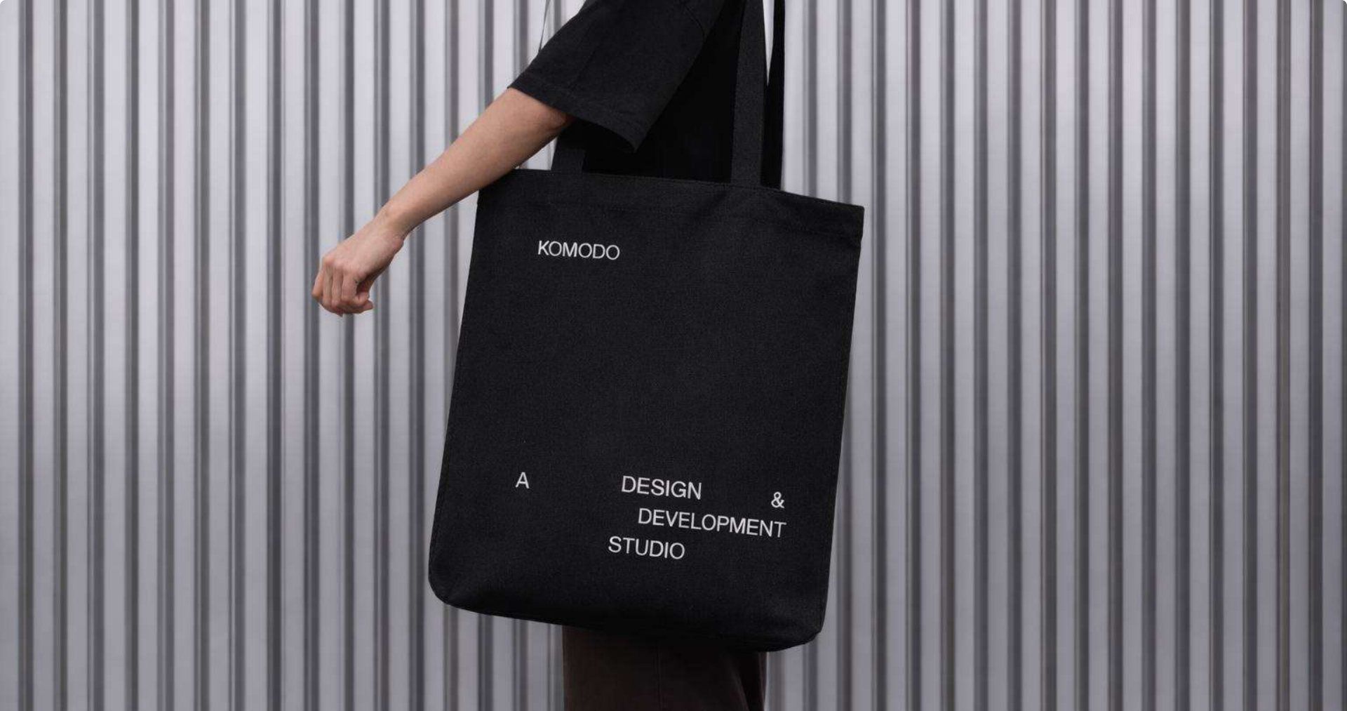 A black Komodo tote bag printed with the wordmark and studio line, held against a corrugated metal wall.